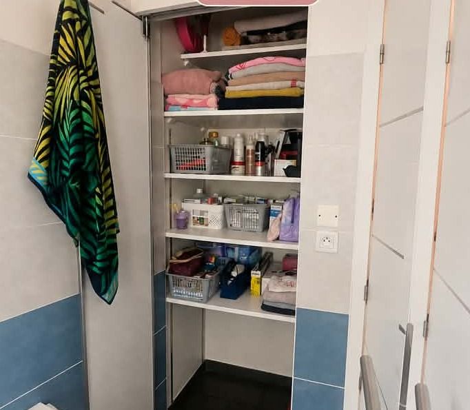 Le même placard réorganisé : linge plié et empilé par couleur en haut, produits regroupés dans des paniers sur les étagères basses