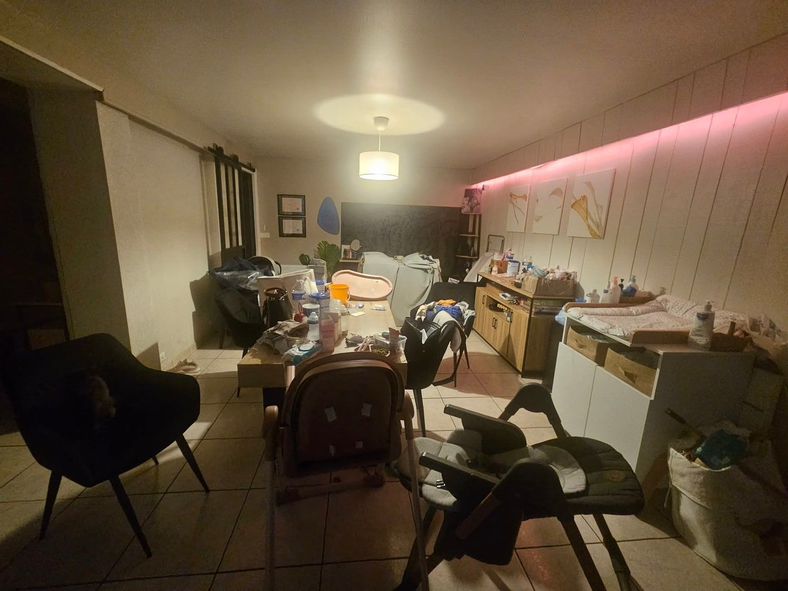 Salle à manger dont la grande table disparaît sous les objets, chaises repoussées en désordre et plan de travail encombré au fond
